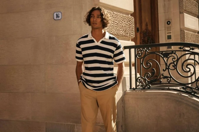 A young adult man in a striped polo shirt stands outside a building with a wrought-iron railing on a sunny day, looking away.