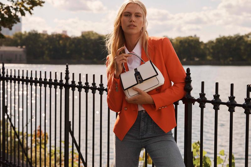 A woman in an orange blazer stands by a fence holding a white handbag near a body of water with city buildings in the background.