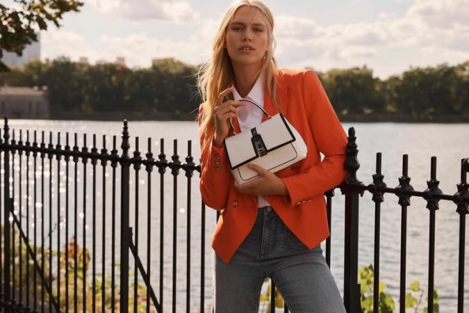 A woman in an orange blazer stands by a fence holding a white handbag near a body of water with city buildings in the background.