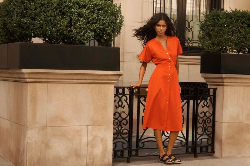Elegant adult female in a button-down orange midi dress posing outdoors against a stone building with wrought iron fencing and topiary.