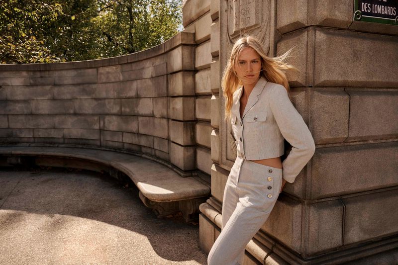 An adult woman with long blonde hair poses in a cropped blazer and pants in front of a stone wall on a sunny day.