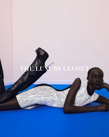 Editorial fashion shot of a Black female model in a white pattern dress, reclining on a blue surface, wearing high, black, leather boots.