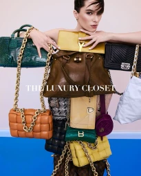 Model holding and surrounded by numerous luxury designer handbags including green, brown, tan, yellow, and black styles.