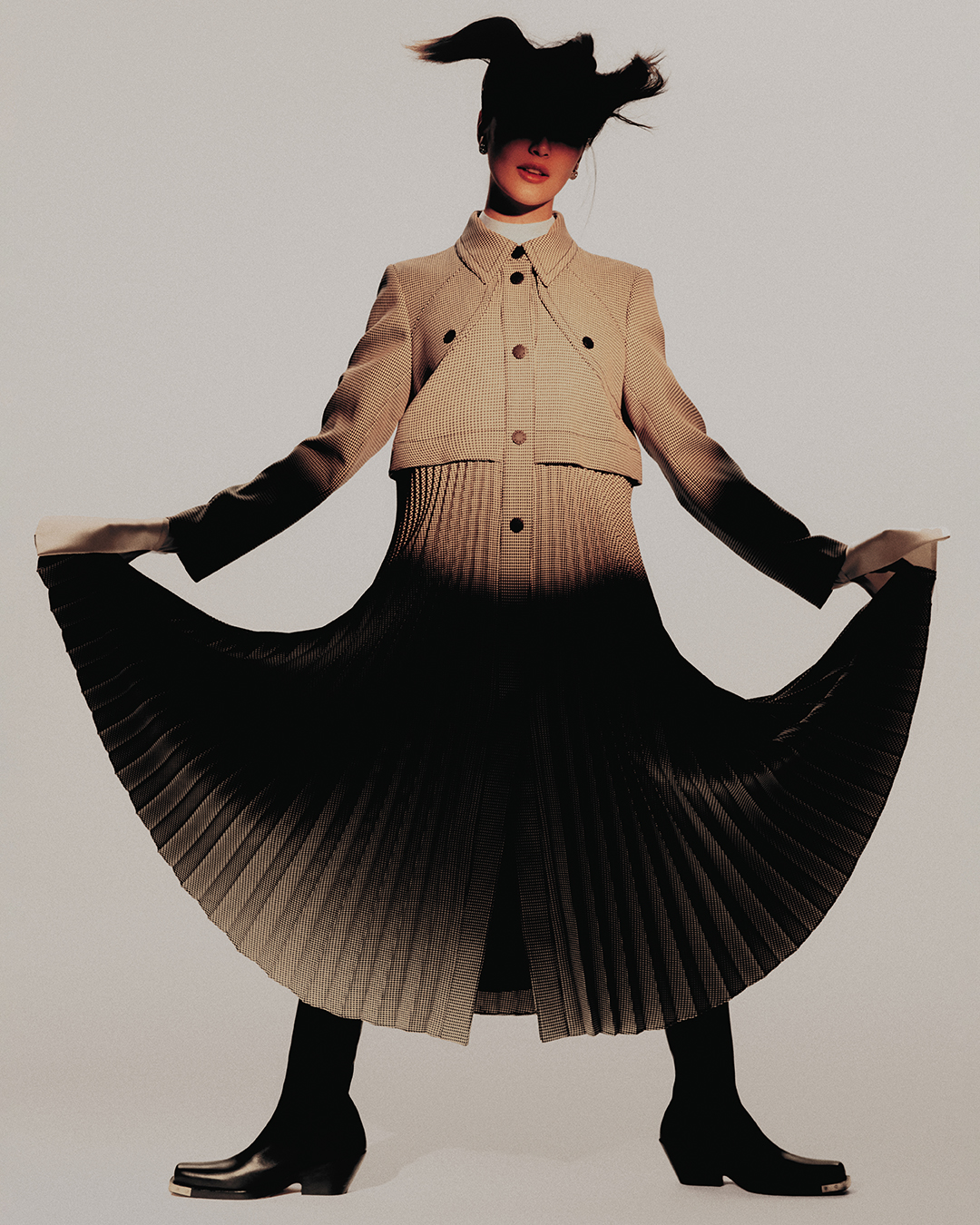 Fashionable adult woman in a beige jacket, pleated skirt, and black boots strikes a pose in a light studio with wind-blown hair.