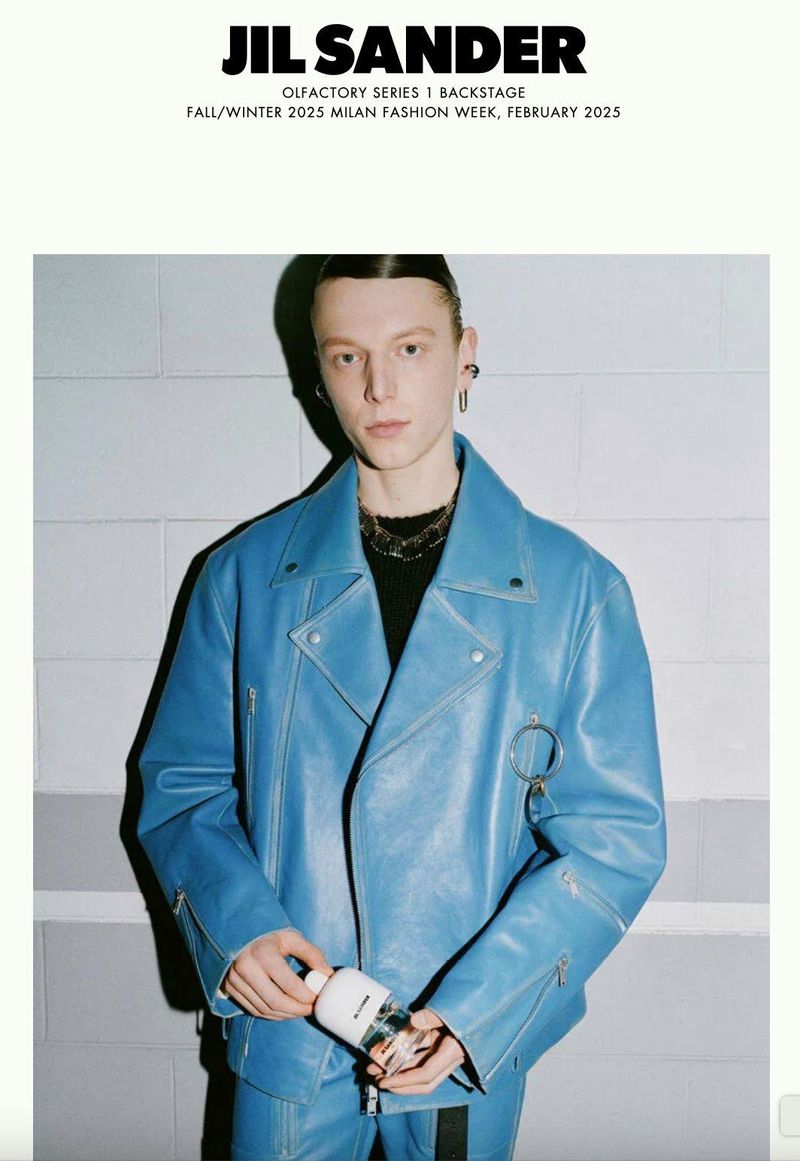 Runway fashion: a fair-skinned male model showcases a light blue leather motorcycle jacket and pants, holding a white perfume bottle, indoors.