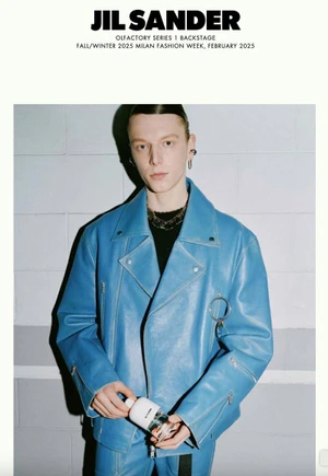 Model wearing a blue leather jacket holding a white perfume bottle against a tiled wall for Jil Sander.