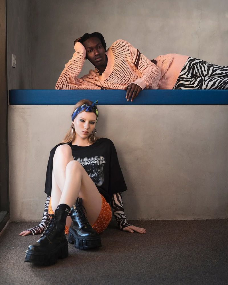 Two fashion models pose indoors. One woman is sitting in the foreground and a man is lying on a blue ledge in the background.