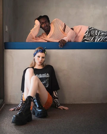 Two fashion models pose indoors. One woman is sitting in the foreground and a man is lying on a blue ledge in the background.