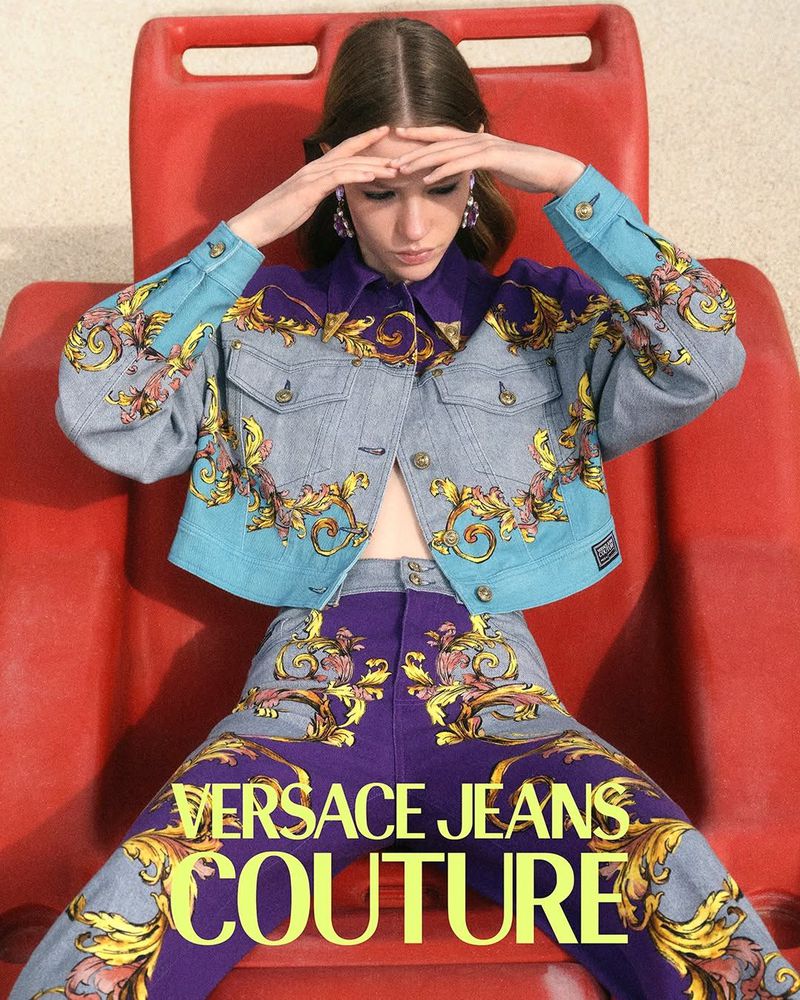 A stylish woman in a Versace Jeans Couture outfit, posing in a red chair, showcasing fashion and modern style with luxury apparel.