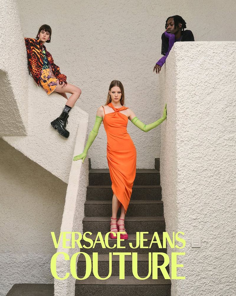 Three fashion models showcase Versace Jeans Couture against a neutral staircase backdrop, featuring dresses and bold color contrasts.