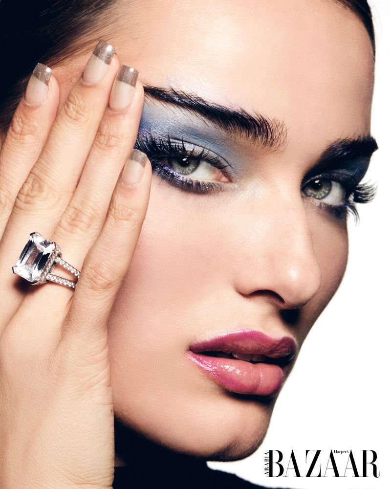 Close-up fashion shot of an adult woman with smokey blue eyeshadow, glossy pink lipstick, and large diamond ring posing in studio.