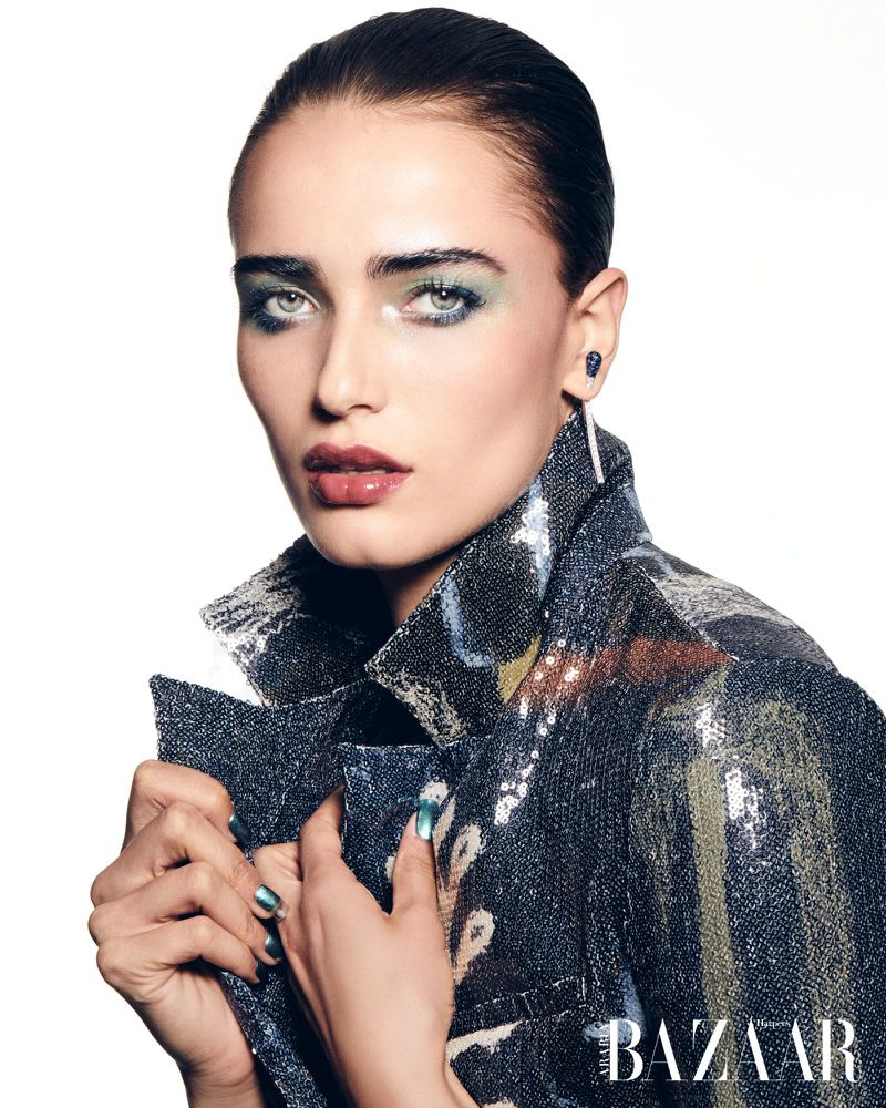Close-up of a female fashion model with blue-green eyeshadow, dark lip color, and a sparkly, patterned jacket in a studio shot.