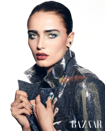 Close-up of a female fashion model with blue-green eyeshadow, dark lip color, and a sparkly, patterned jacket in a studio shot.