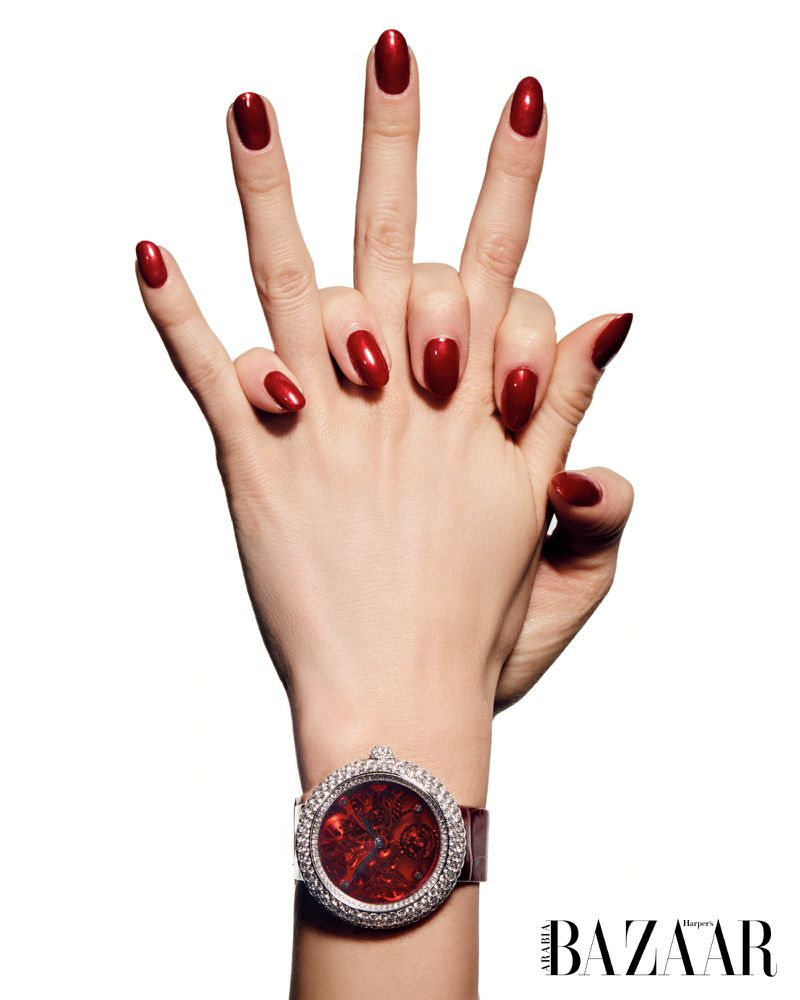 Two hands with red manicured nails overlap, one wrist wearing a red and diamond watch, set against a white studio background.