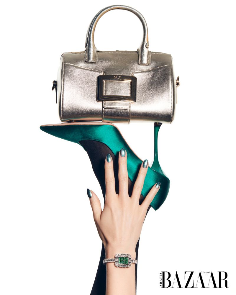 Still life of a silver metallic handbag balanced on a green stiletto heel held up by a woman's hand wearing an emerald bracelet.