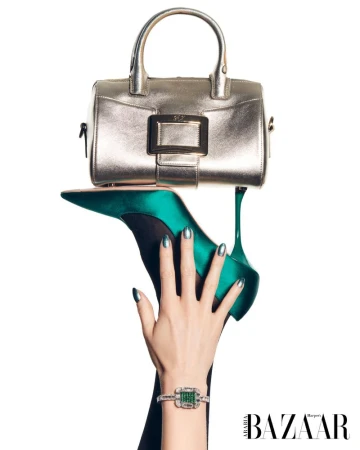 Still life of a silver metallic handbag balanced on a green stiletto heel held up by a woman's hand wearing an emerald bracelet.