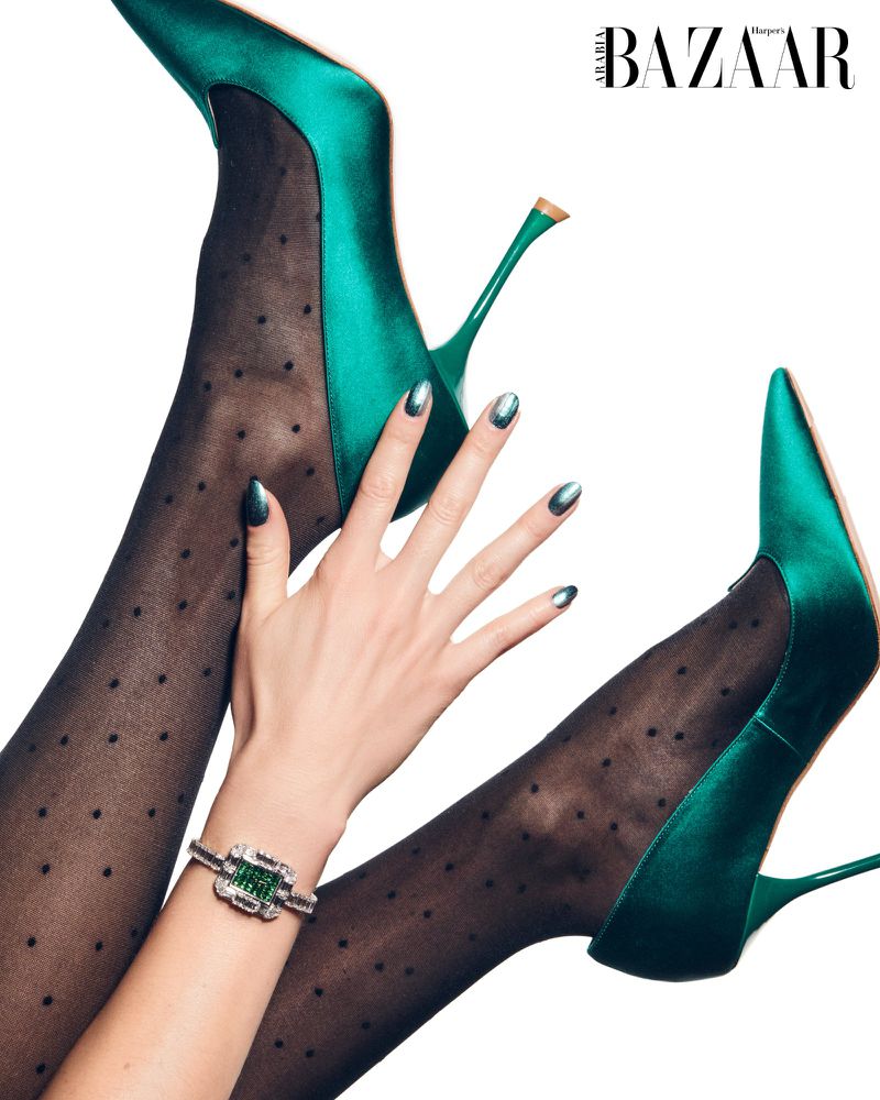 Fashion editorial shot of legs in dotted tights and green satin heels with a woman's hand wearing a green gemstone bracelet.