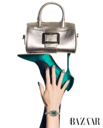 A hand wearing a bracelet and rings holds up a green high-heeled shoe which supports a metallic handbag against a white background.
