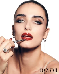 Close-up portrait of a woman applying red lipstick with a brush wearing drop earrings and a diamond ring against a white background