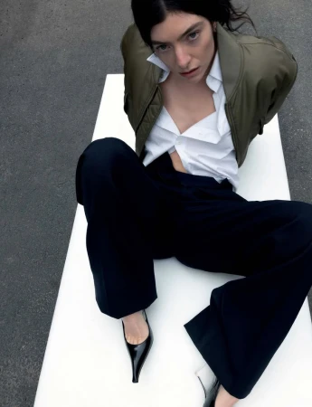 A woman with dark hair sits on a white surface wearing a dark green jacket, open white shirt, dark pants, and black heels.