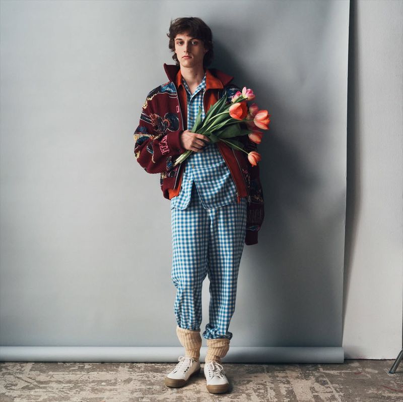 Full-length studio shot of a young adult male in pajama-style outfit holding pink tulips against a neutral gray background.