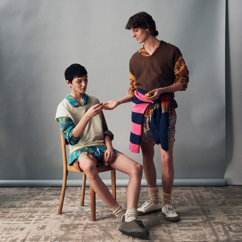 Two adult male models pose indoors on patterned flooring. One sits in a chair handing a small fruit to the other man standing.
