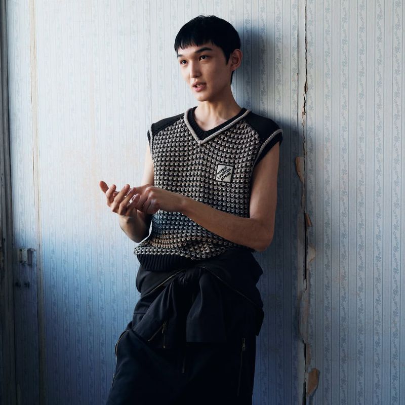 Young adult poses indoors wearing a knit vest and black pants in a fashion shot, near a peeling blue wallpaper background.