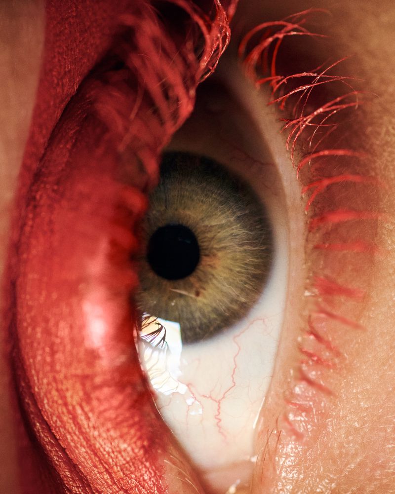 Extreme close-up shot of a human eye featuring green iris, black pupil, red blood vessels, and red eyelashes and brow.