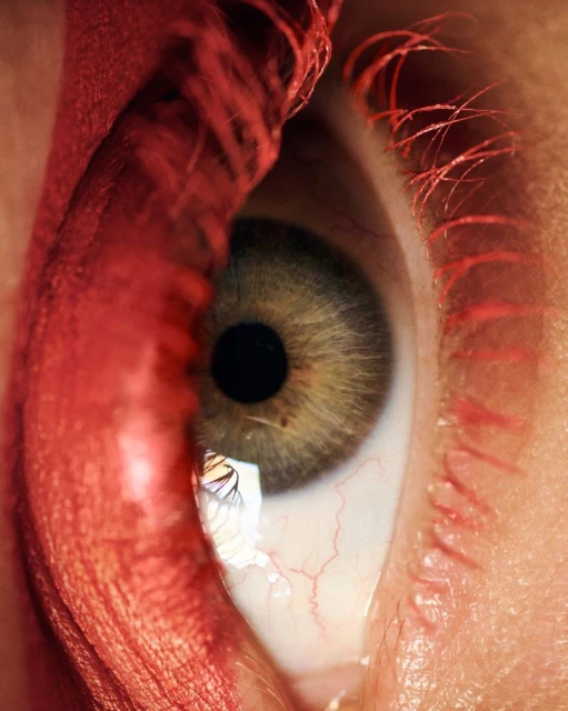 Extreme close-up shot of a human eye featuring green iris, black pupil, red blood vessels, and red eyelashes and brow.