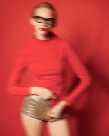 Blurred fashion shot of a woman in a red top and gold sequin shorts against a red backdrop, wearing glasses and red lipstick.