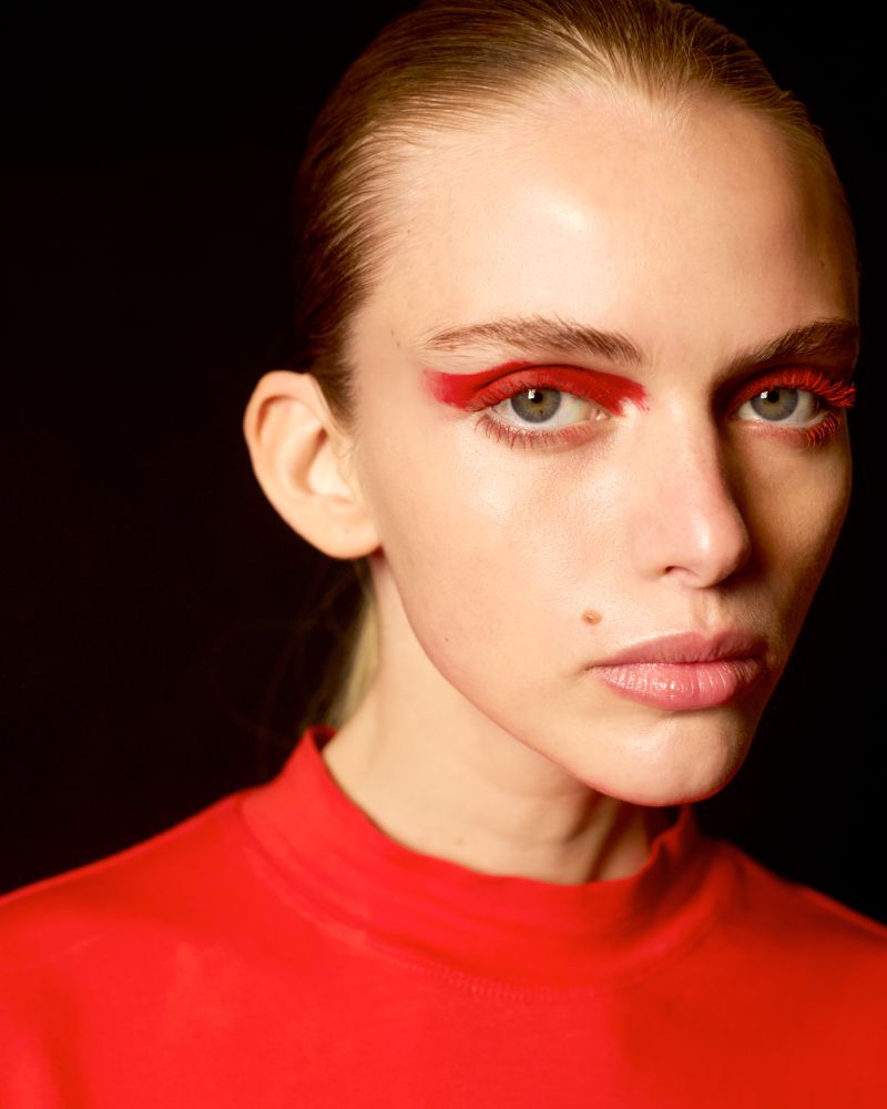 Close-up portrait of a young adult female with red eye makeup and slicked-back blonde hair in a simple red top against a black backdrop.