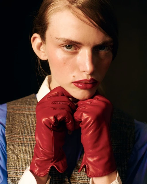 Portrait of a young adult woman in red gloves, with red lips and fair skin, wearing a blue collared shirt and plaid vest.
