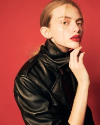 Model in black leather jacket with red lipstick resting chin on hand against a solid red background for Schon Magazine editorial.