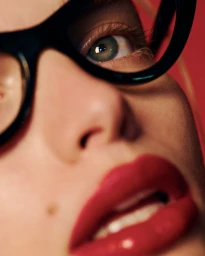 Close-up portrait of a woman wearing thick black glasses and red lipstick against a red background.