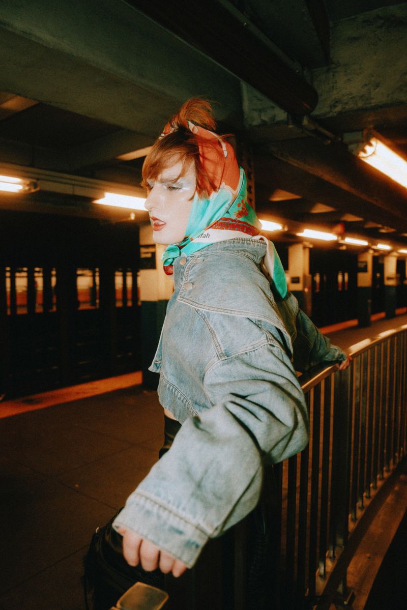 Woman in blue denim jacket and patterned headscarf poses in a subway station with dim lighting and a metal railing, looking away.