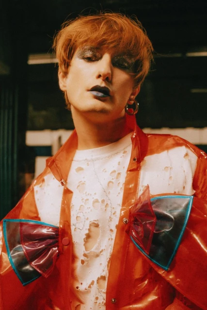 A person with orange hair, wearing a transparent orange jacket with decorative bows, stands indoors against a dark, out-of-focus background.