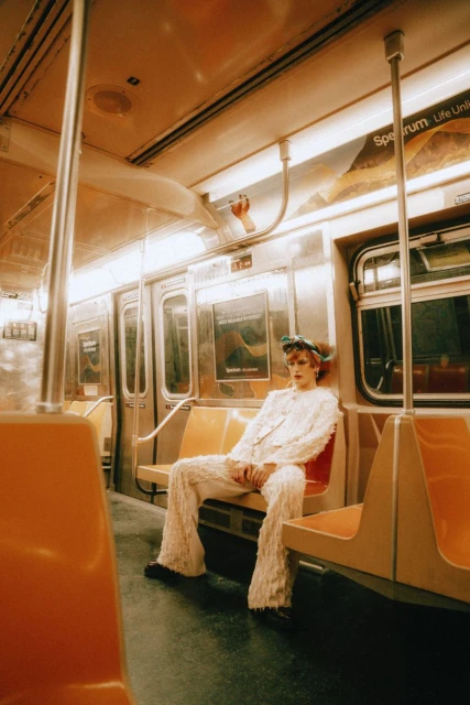 A person in a white suit sits inside a subway car with orange seats, next to the train window under the light with muted orange tone.
