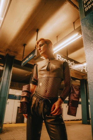 Person poses in an underground subway station wearing an avant-garde leather top, fishnet shirt, and leather pants with metallic arm cuffs.
