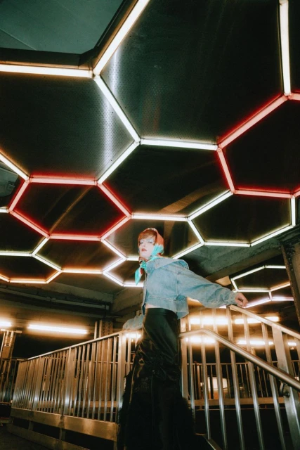 A low-angle shot of a woman on stairs in denim jacket and long black skirt, with a modern lighting fixture on the ceiling.