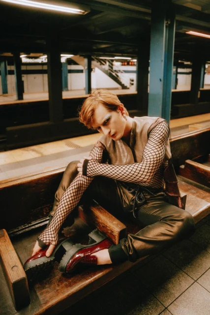 Non-binary person with red hair poses on a wooden bench in a subway station wearing a vest, fishnet top, black pants, and patent-leather shoes.