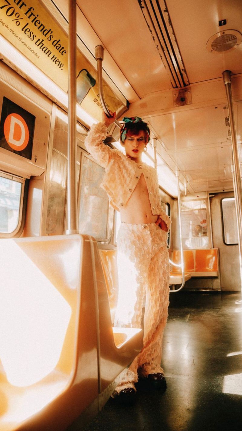Young adult poses in an open lace outfit on a subway train with yellow seating and interior details, bathed in bright, diffused lighting.