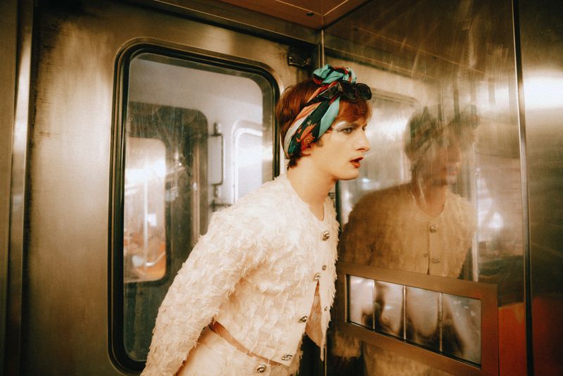 A person with reddish hair and a headscarf in an off-white jacket and skirt posing inside of a metal train car.