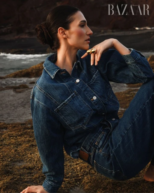 Brunette woman in denim jacket and jeans poses on a rocky shore with crashing waves in the background for a fashion editorial.