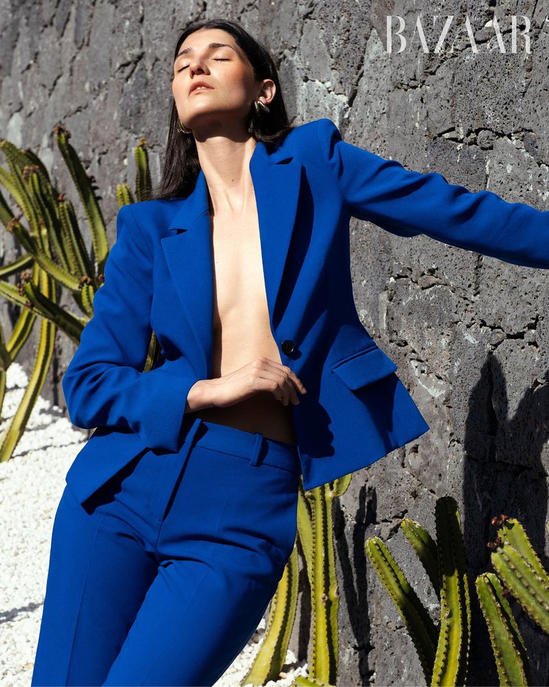 Woman in a blue suit poses against a textured wall with cacti; fashion editorial style, natural light, BAZAAR logo visible, full shot.