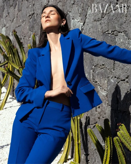 Woman in a blue suit poses against a textured wall with cacti; fashion editorial style, natural light, BAZAAR logo visible, full shot.
