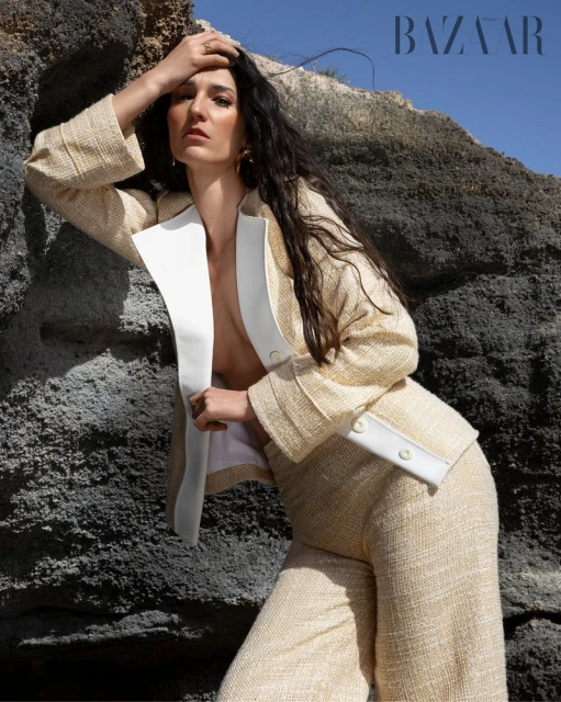A woman poses in a cream-colored, tweed pantsuit against a rocky landscape, wearing a blazer with white lapels, shot in natural light.