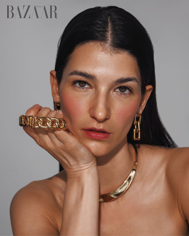 Close-up portrait of a woman with fair skin wearing a gold necklace, earrings, and statement ring against a neutral backdrop.