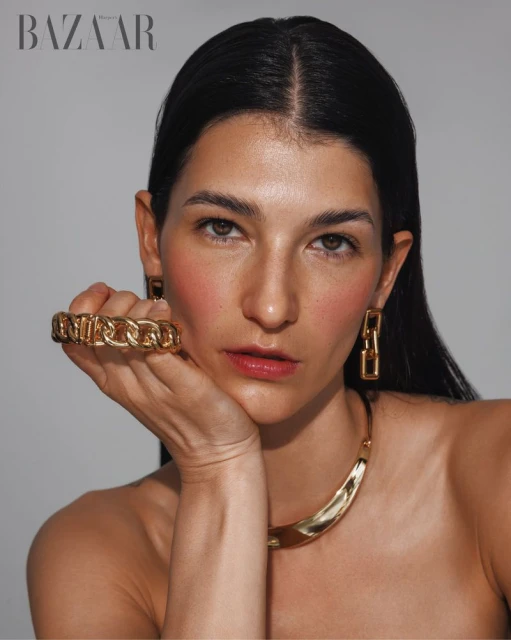 Close-up portrait of a woman with fair skin wearing a gold necklace, earrings, and statement ring against a neutral backdrop.