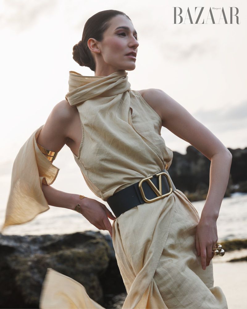 A fashion model poses outdoors in a draped cream dress with a black belt and gold accents, standing on rocks by a beach with BAZAAR logo.