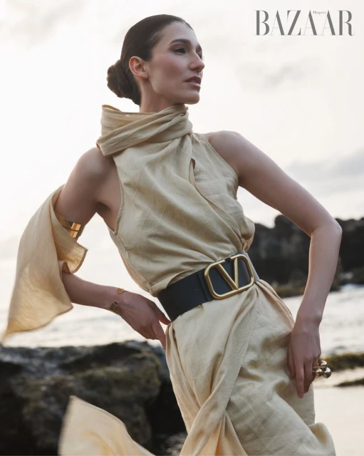 A fashion model poses outdoors in a draped cream dress with a black belt and gold accents, standing on rocks by a beach with BAZAAR logo.